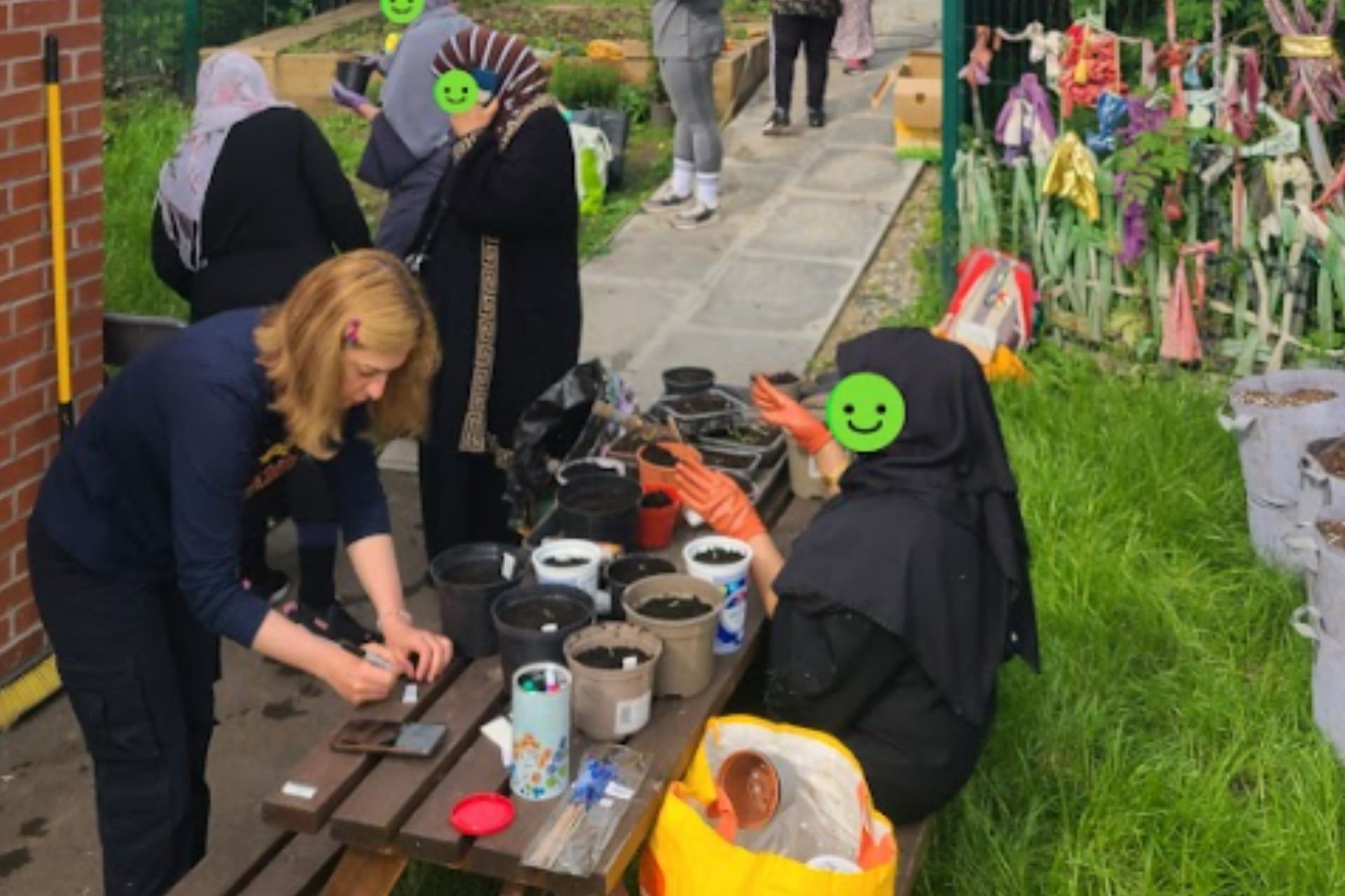 An ethnically diverse group of women gather around a table. On the table there are plant pots filled with soil as the women are placing seeds and plant cuttings into them.