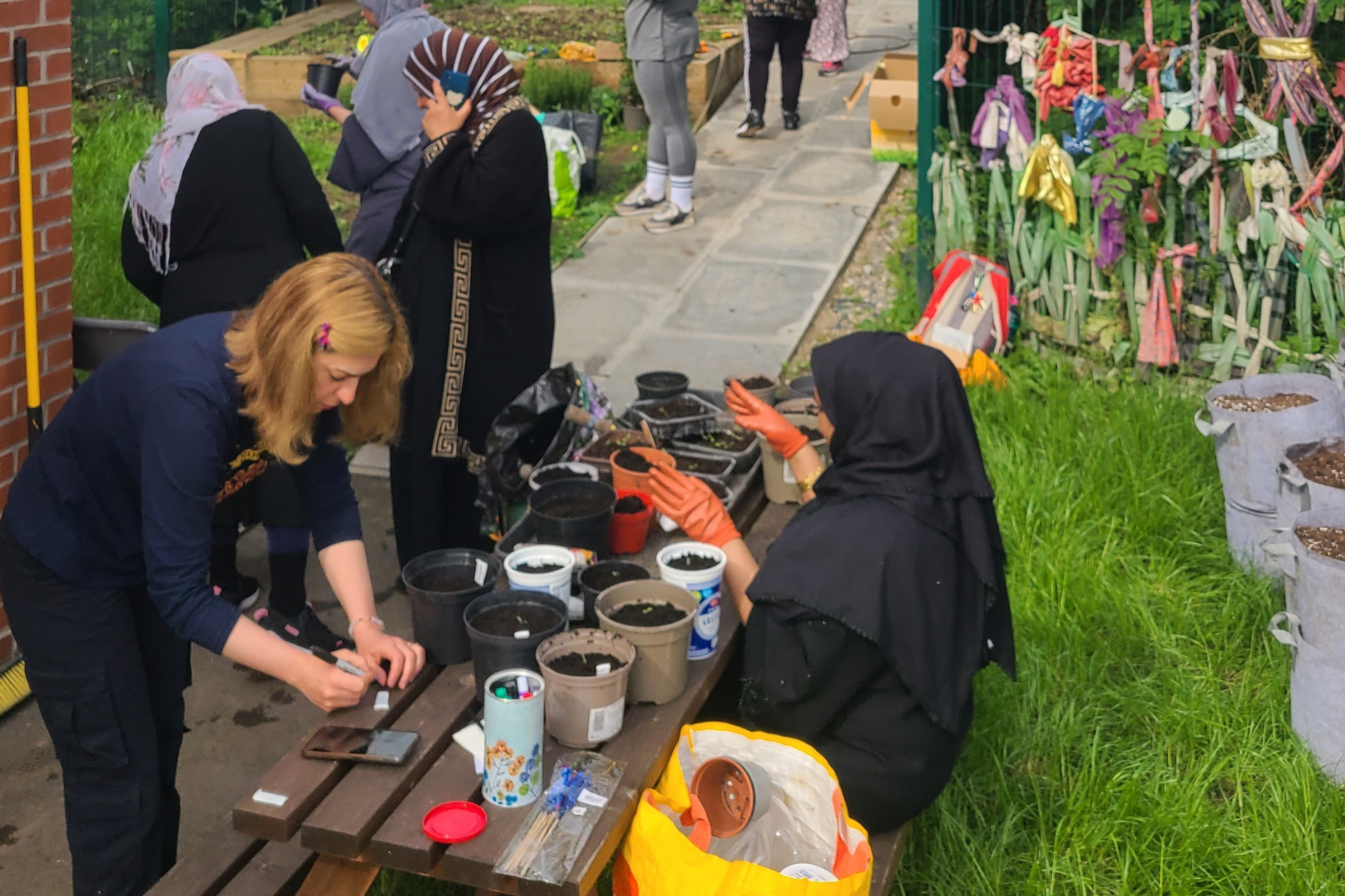 An ethnically diverse group of women gather around a table. On the table there are plant pots filled with soil as the women are placing seeds and plant cuttings into them.