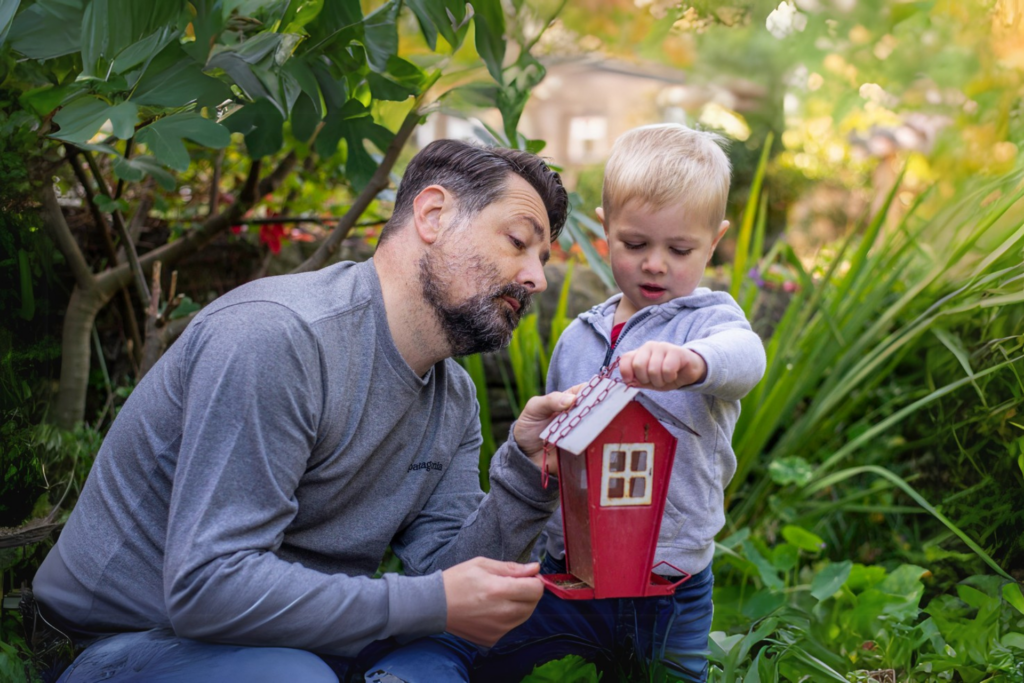 A man squatting down shows a red bird house he is holding to a young boy.