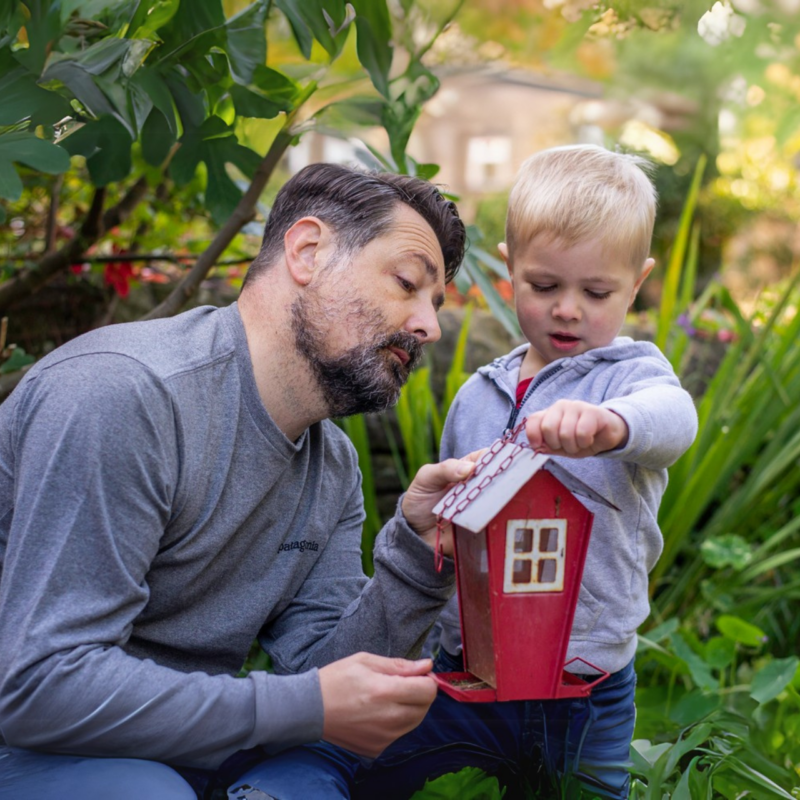A man squatting down shows a red bird house he is holding to a young boy.