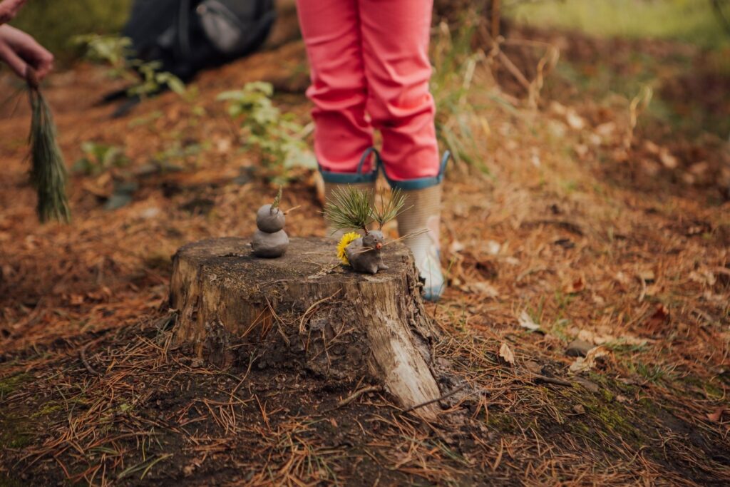 Image shows a child wering wellies in a woodland, standing behind a piece of wild art they have made with natural materials