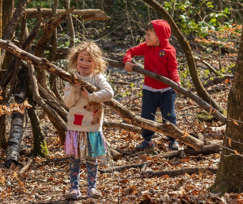 Two children playing in the woods. The little girl in front is carrying a giant branch and looks very happy